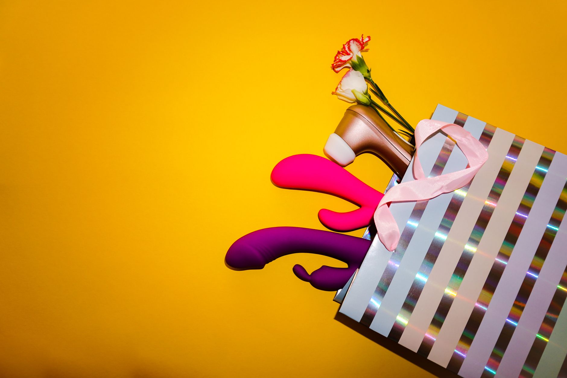 Vibradores coloridos con flores en una bolsa de regalo holográfica sobre fondo amarillo brillante